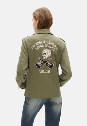 Chaqueta de estilo militar en verde oliva con un parche de calavera y guitarras cruzadas en la parte posterior, que presenta texto bordado y un corte relajado.