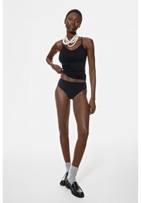 Zwarte tanktop en high-waisted bikini. Model draagt zwarte schoenen, grijze sokken en een gelaagde parelketting op een witte achtergrond.