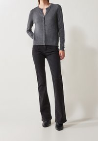 Kobieta w szarej, zapinanej kardiganie, czarnych dzwonowatych dżinsach i czarnych botkach na obcasie stojąca na jednolitym jasnym tle.