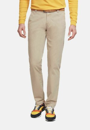 Mann trägt beige Hose, ein gelbes Langarmshirt, einen braunen Gürtel und schwarze, gelbe, orange und weiße Sneakers.