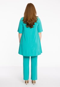 Een turquoise tuniek met korte mouwen, gecombineerd met bijpassende broeken, met een ontspannen pasvorm en een gladde textuur, bekeken vanaf de achterkant.