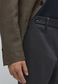 Bruine gestructureerde blazer met zwarte knopen, gedragen over een marineblauwe shirt, gecombineerd met donkergrijze broek met knoopsluiting en gladde stof.