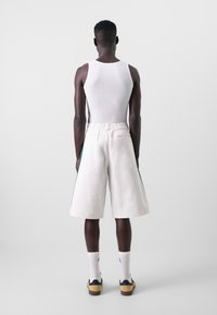 Man som står med ryggen mot kameran, klädd i en vit linne, vita vida shorts med sidostrippar, vita strumpor och gula sneakers.