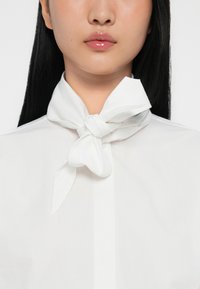 Camicia bianca con una sciarpa di seta legata al collo, texture liscia e design pulito, che mette in risalto un'estetica minimalista.