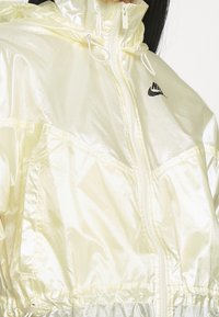 Gul iriserande vindjacka med hög krage, dragkedja och svart Nike-logotyp. Har elastiska ärmslut och en samlad nederkant för en åtsittande passform.