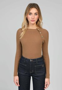 Haut marron à manches longues côtelé avec un col arrondi et des boutons décoratifs sur l'épaule gauche, associé à un jean en denim foncé taille haute.