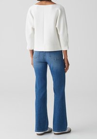 Pull blanc texturé à encolure large, assorti à un jean évasé bleu. La tenue présente une coupe décontractée et des détails de coutures discrètes.