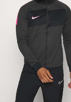 Uomo che indossa una giacca sportiva grigio scuro e nera con logo Nike rosa e pantaloni neri con logo Nike bianco, in piedi davanti a uno sfondo semplice.