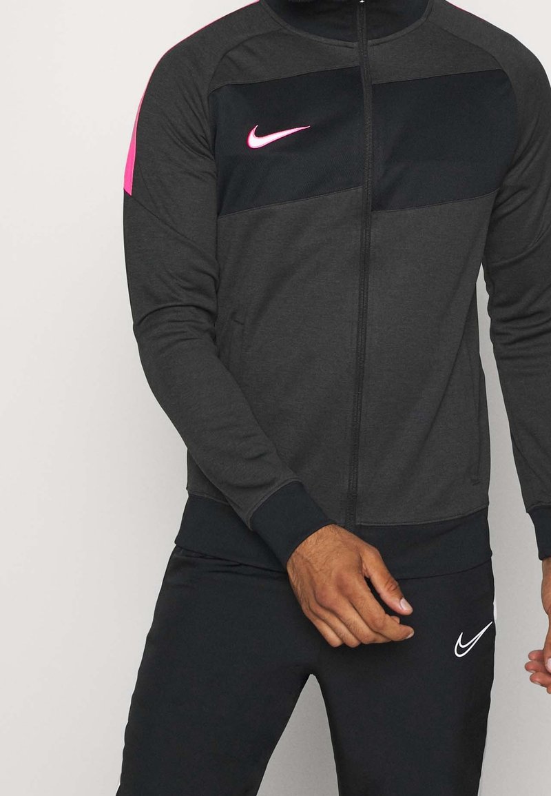 Mann in dunkelgrau und schwarzer Sportjacke mit pinkem Nike-Logo und schwarzen Hosen mit weißem Nike-Logo, steht vor einfarbigem Hintergrund.