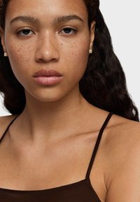 Zbliżenie osoby o średnim odcieniu skóry, widoczne piegi, ciemne falowane włosy, jasne błyszczące usta i małe perełkowe kolczyki w sztyfcie, ubrana w brązową bluzkę.