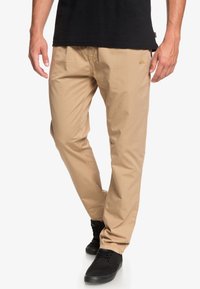 Beige avslappnade byxor med en ledig passform, med synliga stygn och en liten logga på framfickan, parat med svarta sneakers.