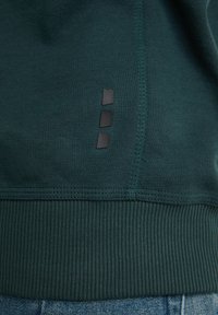 Orlo di felpa verde scuro con tre piccole patch rettangolari nere in gomma sopra il polsino a coste, indossata sopra jeans di denim blu.