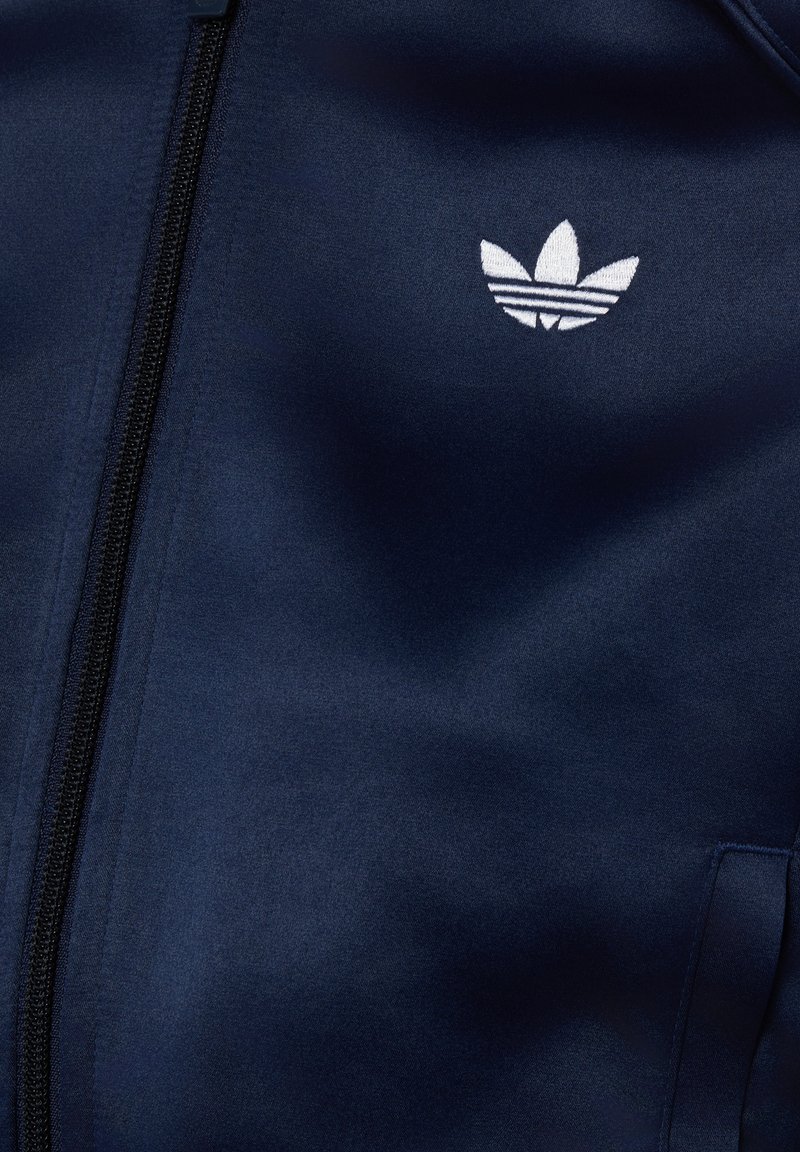 Navyblaue Jacke mit sichtbarem Frontreißverschluss und weiß gesticktem Adidas Trefoil-Logo auf der Brust nahe der Tasche.