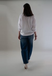 Vrouw die wegloopt in een witte blouse, blauwe jeans, beige hakken en een armband om haar rechterpols tegen een plain achtergrond.