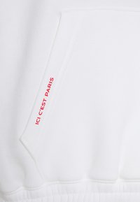 Sudadera blanca con una textura suave que presenta un bolsillo frontal y el texto rojo "ICI C'EST PARIS" en un lateral, mostrando un diseño casual.