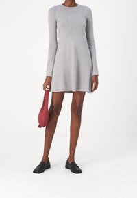 Donna che indossa un vestito corto a maniche lunghe grigio chiaro, tiene in mano una piccola borsa rossa e indossa mocassini neri, mentre si trova su un pavimento bianco.