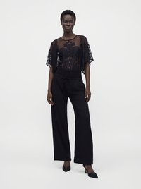 Femme debout portant un haut en dentelle noire transparente à motif floral, un pantalon noir à jambes larges et des chaussures à talons noirs sur fond blanc.