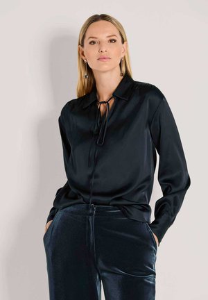 Zwarte satijnen blouse met een strik-hals, lange mouwen en een losse pasvorm. Gecombineerd met glanzende, donkerblauwe fluwelen broek.