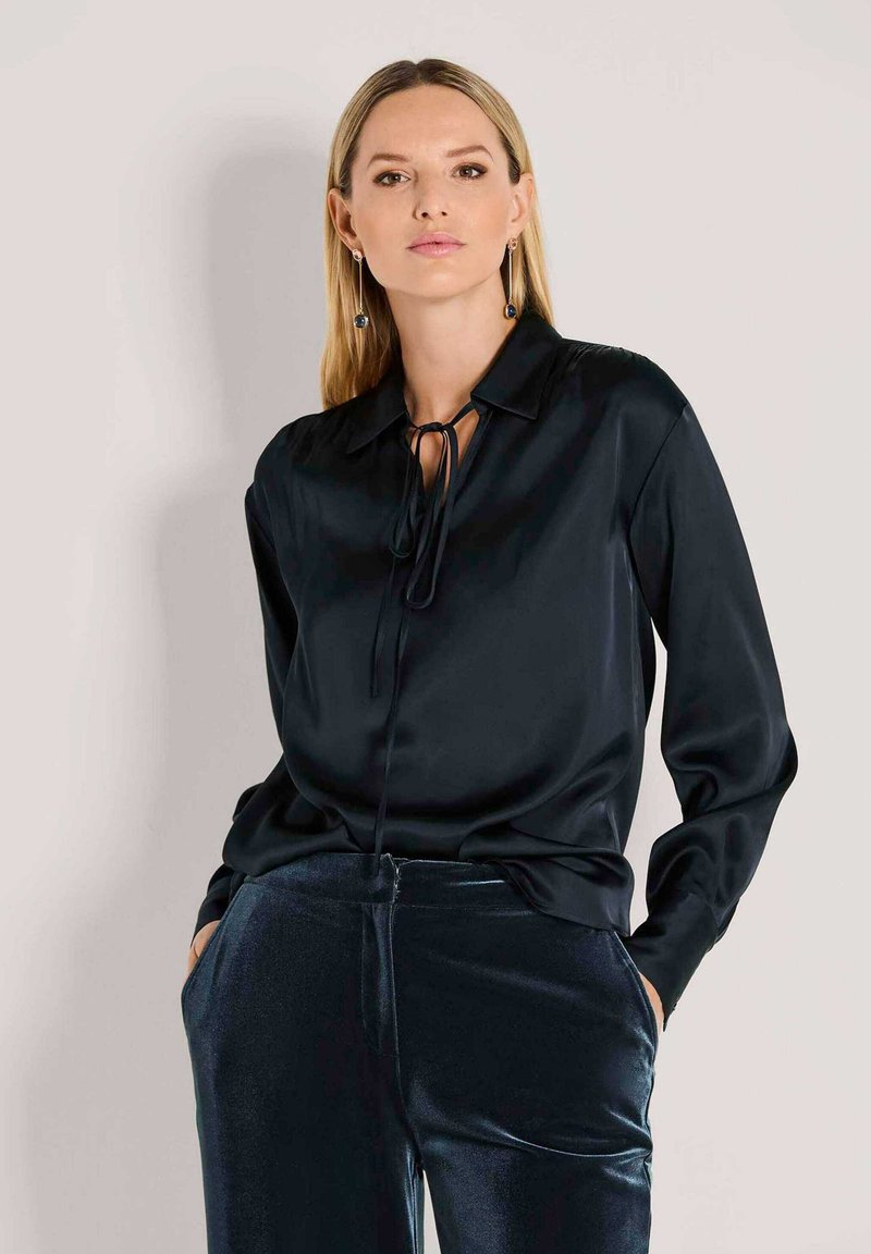 Schwarze Satinbluse mit Bindegürtel, langen Ärmeln und lockerer Passform. Kombiniert mit schimmernden, dunkelblauen Samenhosen.