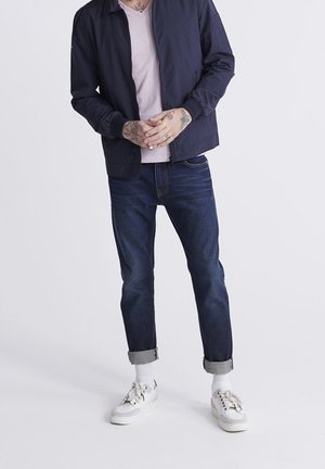 Person iført en marineblå jakke, lyserød T-shirt, opsmøgede mørke jeans, hvide sokker og hvide sneakers, står med hænderne foldet.