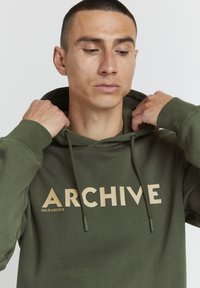 Oliven farvet hoodie med et stort beige "ARCHIVE" print, snoretræk og ribbede manchetter, vist mod en ensfarvet baggrund.