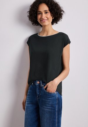 Donna con capelli ricci che sorride, indossa una maglietta nera a maniche corte e jeans blu, in piedi contro un muro chiaro e semplice.