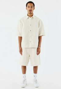 Lichtbeige kortemouwshirt met een subtiele textuurpatroon, knoopsluiting, gecombineerd met bijpassende shorts. Witte sneakers en crew-sokken maken de look compleet.
