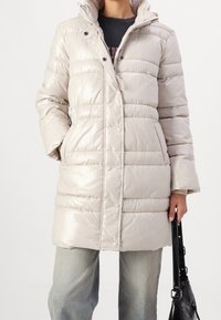 Osoba v lehké béžové prošívané bundě se zapínáním na druky, v kombinaci s světle modrými džínami, držící černou kabelku po boku.