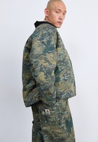 Giacca a motivo camouflage realizzata in tessuto resistente, con vestibilità rilassata, tonalità verde e marrone, e tasche laterali.