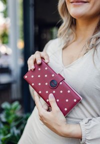 Red leather wallet with white polka dots, rectangular shape, secured with a black button clasp. Smooth texture with a glossy finish.