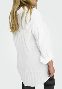 Donna con lunghi capelli biondi che indossa una blusa bianca plissettata con maniche a tre quarti e pantaloni neri, voltata verso l'indietro rispetto alla fotocamera.
