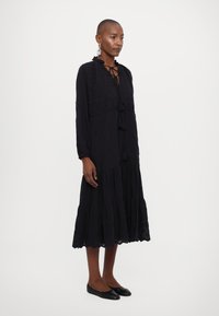 Vestido preto de mangas longas com bordados florais, cintura franzida e bainha em camadas. Apresenta laços no pescoço com franjas. Usado com sapatos pretos de sola plana.