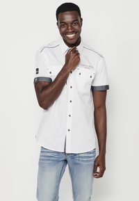Camisa blanca de manga corta con puños acentuados en denim, dos bolsillos en el pecho y cierre de botones. Presenta un logo en la manga y costuras decorativas.