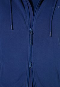 Giacca sportiva blu Navy con zip, dal tessuto liscio, dotata di zip frontale, cappuccio regolabile con lacci e branding discreto sul petto.