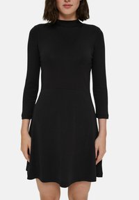 Svart klänning med hög halsringning och trekvart ärmar, med en figurnära bodice och utsvängd kjol. Tillverkad av smidig, elastisk tyg.