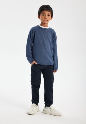 Pull en tricot bleu avec col rond, associé à un pantalon cargo bleu marine et des baskets blanches. La texture est douce, et le pantalon comporte des poches latérales.