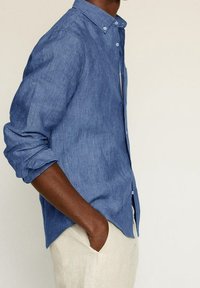 Hellblaues Leinenhemd mit einem Knopfkragen, hochgekrempelten langen Ärmeln und einer Brusttasche. Weiche Textur und lässiges Design.