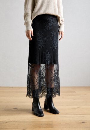 Schwarzer Spitzenrock mit transparentem Saum und Blumenmuster. Weicher Stoff, taillierte Passform, kombiniert mit schwarzen Knöchelstiefeln. Texturierte Oberflächenstruktur.