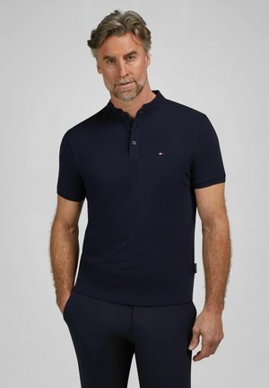 Mężczyzna z siwymi włosami i brodą, ubrany w dopasowaną granatową koszulkę polo z krótkim rękawem oraz granatowe spodnie, na jednolitym tle.