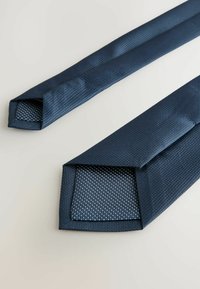 Cravate bleu marine en tissu texturé, avec un extérieur uni et un intérieur à motifs avec de petits points. Les extrémités sont pointues.