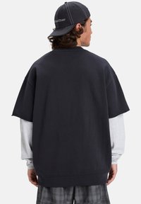 Felpa nera oversize con maniche corte indossata sopra una maglietta grigia a maniche lunghe. Tessuto morbido, design semplice e vestibilità rilassata.