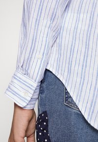 Bílá a modrá pruhovaná košile s dlouhým rukávem, v kombinaci s tmavými džíny z denimu, které mají kapsu s námořnickými tečkami.