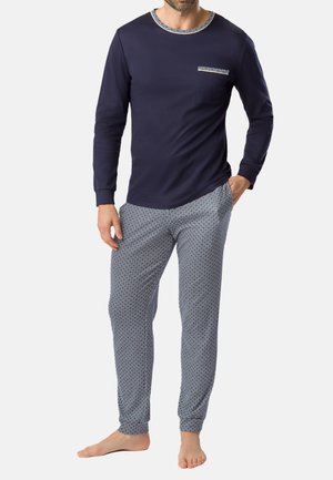 Mann trägt navyblaues Langarmoberteil mit gemustertem Saum und passende gemusterte Pyjamahose, steht barfuß mit einer Hand in der Tasche.