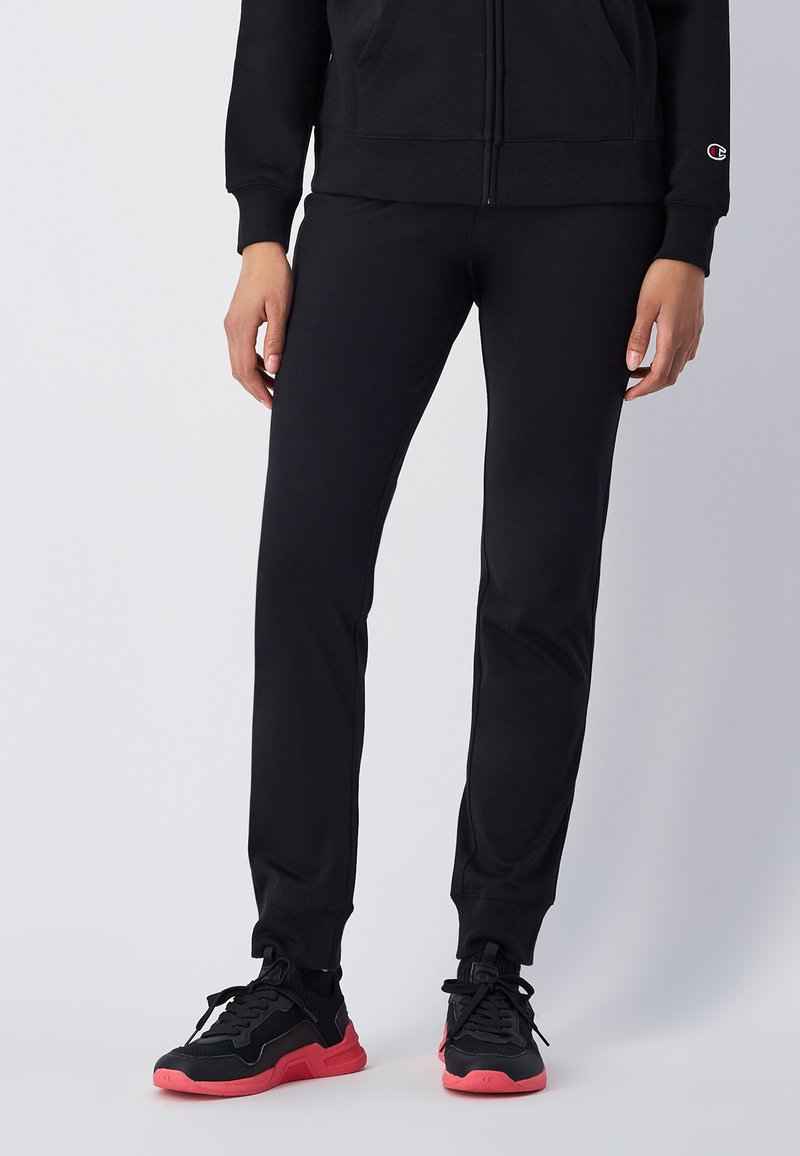 Schwarze Jogginghose aus weichem Stoff, mit schmalem Schnitt, elastischem Bund und gerippten Bündchen. Kombiniert mit schwarzen Sneakers und rosa Akzenten.