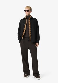 Zwart overhemd, bruine gewatteerde jas, bruine coltrui, losse donkere pantalons en zonnebril. Gladde texturen en neutrale kleuren overheersen.