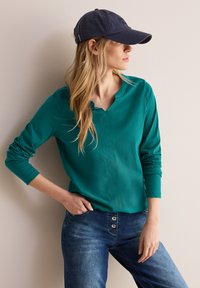 Türkises Langarmoberteil mit einem runden V-Ausschnitt, kombiniert mit dunkellblauen Jeans mit Knopfdetails und einer dunkelblauen Baseballmütze.