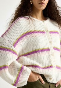 Pletený kardigan v krémové barvě s růžovými, fialovými a zelenými vodorovnými pruhy. Má zapínání na knoflíky a snížená ramena pro volnější střih.