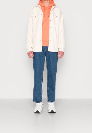 Witte denimjack over een oranje hoodie, blauwe jeans en witte sneakers. Opvallende details zijn de knoopzakken en het contrasterende stiksel.