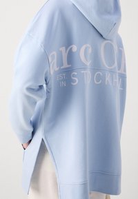 Hellblauer Kapuzenpullover aus weichem Material, mit großem weißem Aufdruck auf der Rückseite und seitlichen Schlitzen als Designdetail.