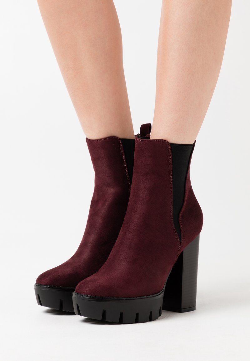 Bottes en daim bordeaux montant à la cheville avec des talons plateformes noirs épais portées sur des jambes nues sur fond blanc.